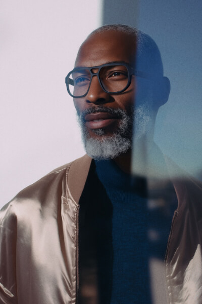 KlenzeBaum Brille Santos Dunkelhäutiger Mann mit goldschimmernder Seidenjacke und graumeliertem Bart trägt eine schwarze Brille