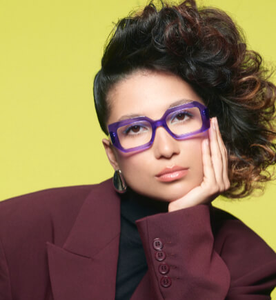 WOOW Brillenmode Wollenhaupt Solingen Portrait einer stilvollen Frau, die den Kopf leicht auf ihre Hand stützt und vor einem leuchtend gelben Hintergrund posiert. Sie trägt eine markante, lila-transparente Brille der Marke WOOW, einen dunkelroten Oversized-Blazer und hat voluminöses, seitlich gestyltes Lockenhaar.