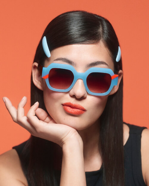 WOOW_SUPER SQUARE_COL1348 - Sonnenbrillen Wollenhaupt Solingen Portrait einer jungen Frau mit dunklem, glattem Haar, hellblauen Haarklammern und orange-rotem Lippenstift vor orangefarbenem Hintergrund. Sie stützt ihr Kinn sanft auf die Hand und trägt eine markante, quadratische Sonnenbrille von WOOW mit hellblauem Rahmen und roten Details an den Seiten.
